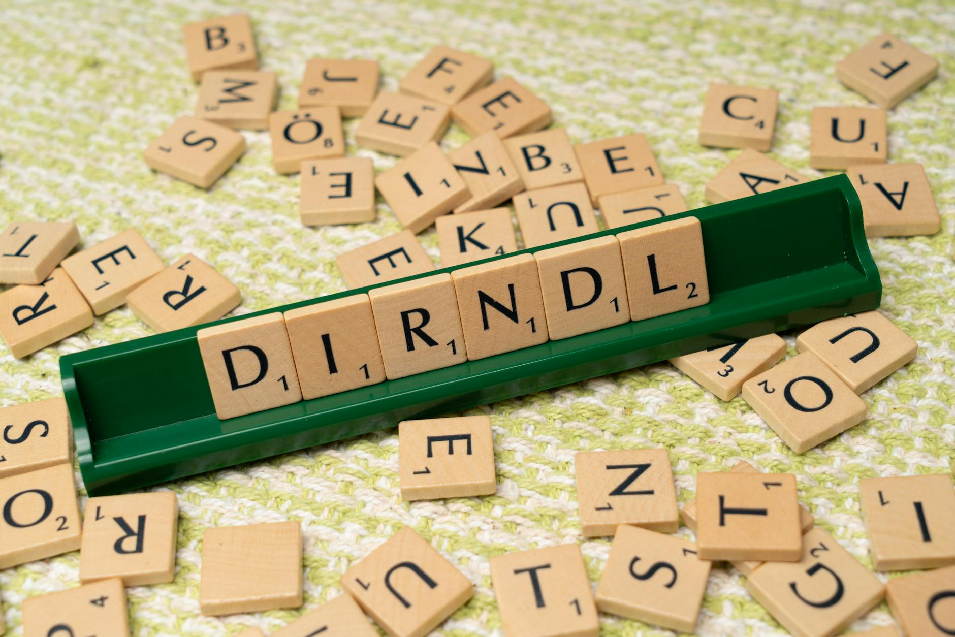 Scrabble tiles spelling 'dirndl' on a green tile holder with scattered letters on a textured surface.